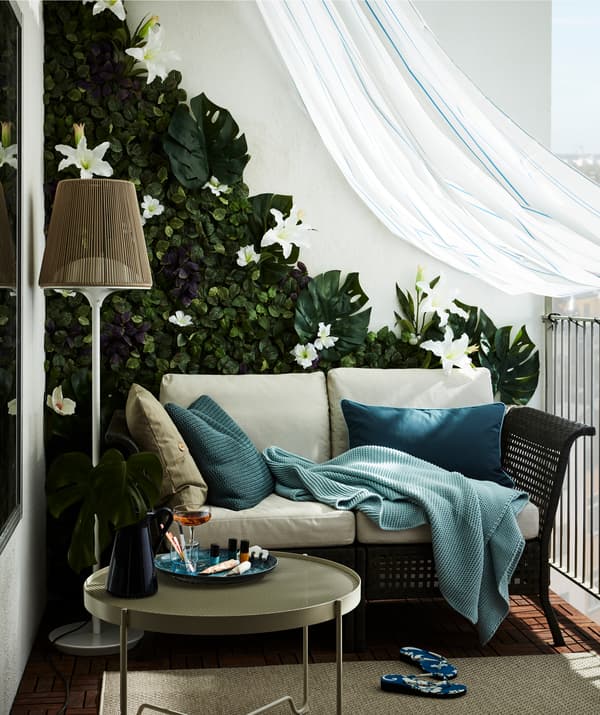 A balcony furnished with a two-seat sofa, artificial-flower wall and draped with a weatherproofing shower curtain.