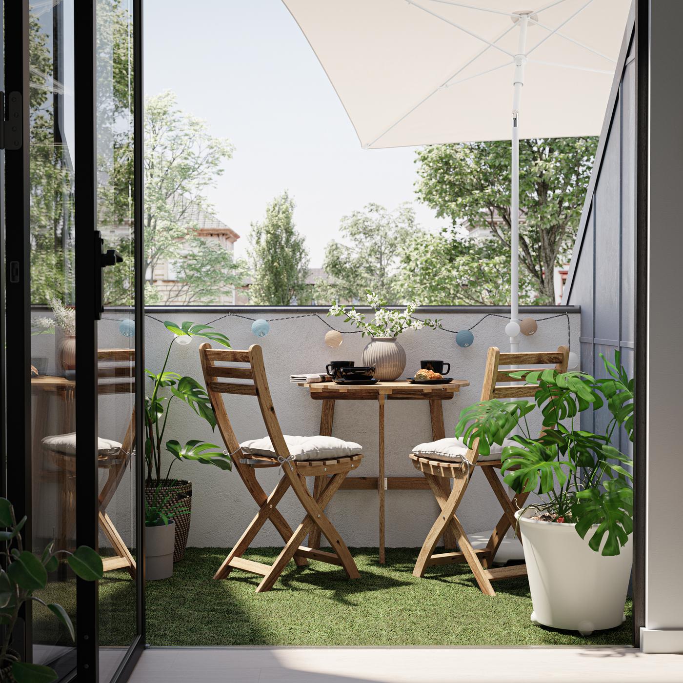 A balcony with two wooden chairs and a table in the ASKHOLMEN series, plus a grey-beige parasol and various potted plants.