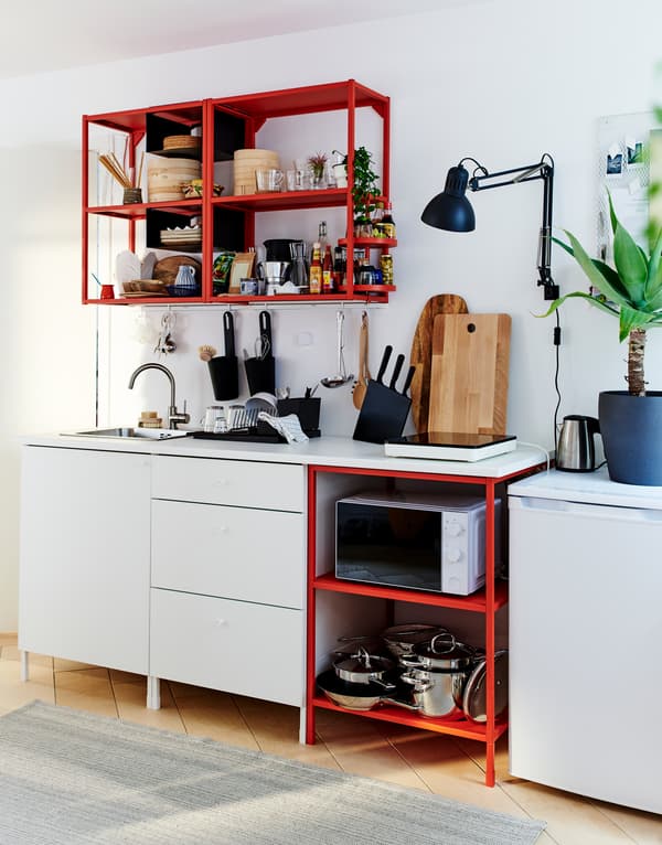 A basic kitchen solution with white drawers, red metal shelves for standing and wall-hung storage, and a low fridge.