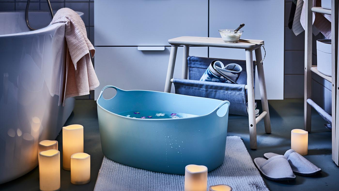 A bathroom mood-lit by scattered LED block candles, a stool placed next to a big footbath, flowers floating in it.