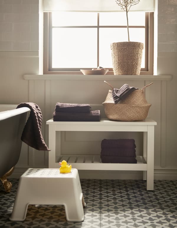 A bathroom with a rattan basket, a white BOLMEN step stool, and a white HEMNES bench with a shelf for towels.