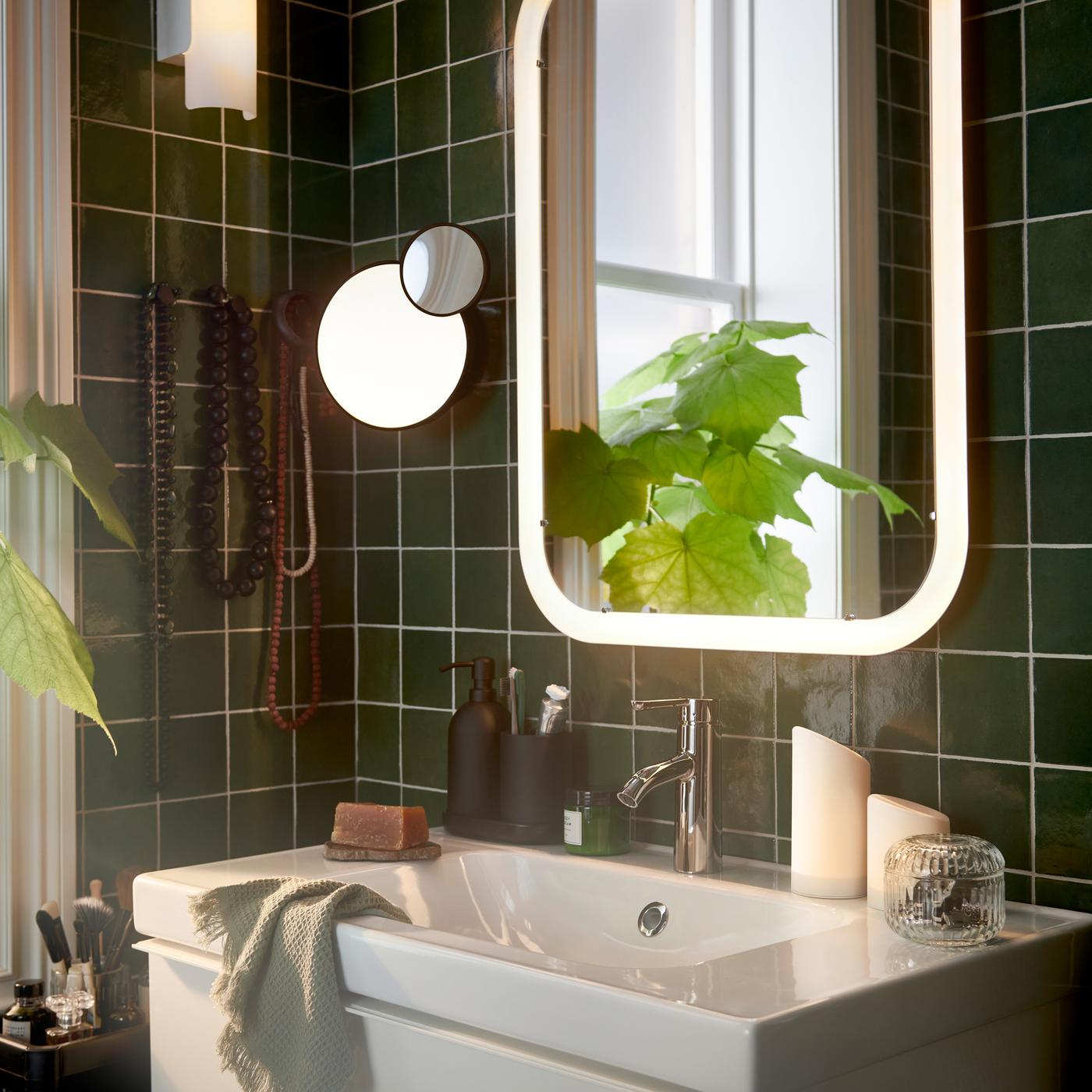 A bathroom with a white mirror with integrated lighting over a wash-stand, plus two LED block candles and a potted plant.