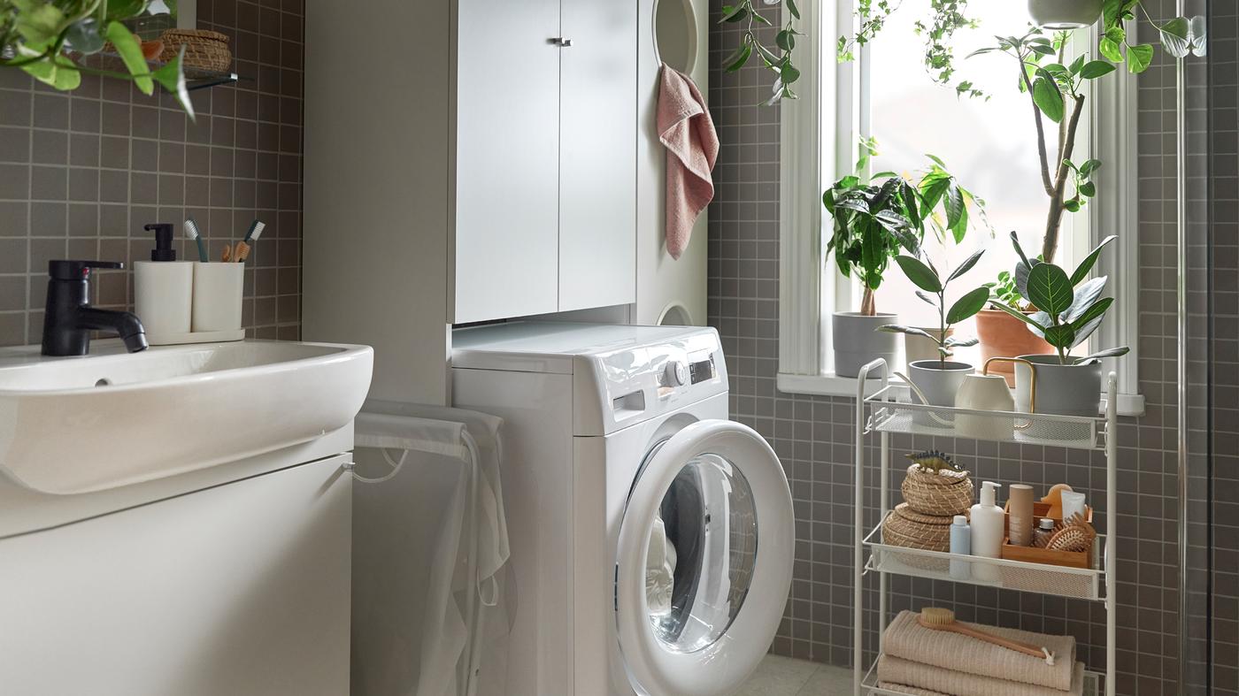 A bathroom with an UDDARP washing machine in white and a HORNAVAN trolley in white with items and a NYSJÖN cabinet in white.