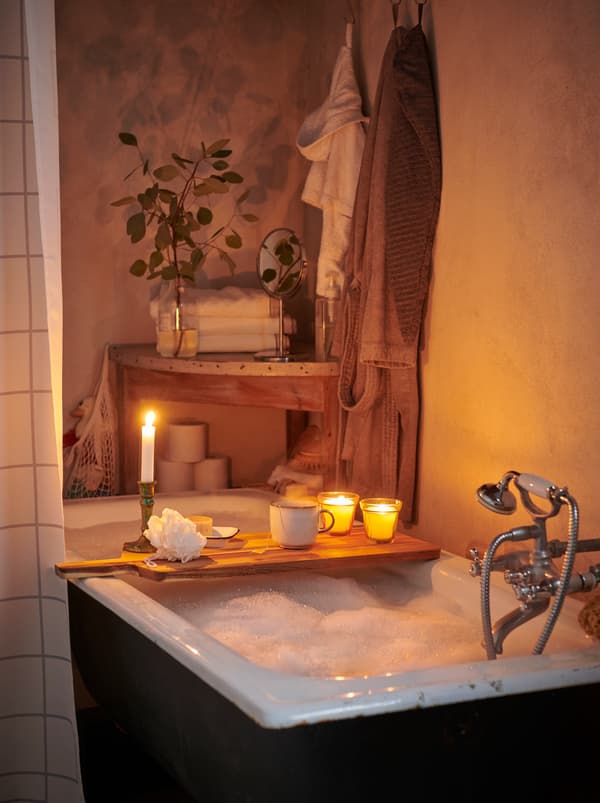 A bathtub full of foamy bubble bath, with a cutting board with candles and other items balanced across the rim.