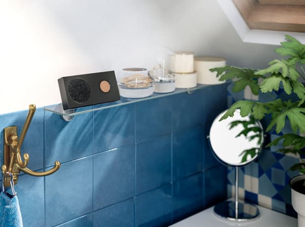 A battery powered IKEA ENEBY black portable Bluetooth speaker on a glass shelf above blue tiles in a bathroom.
