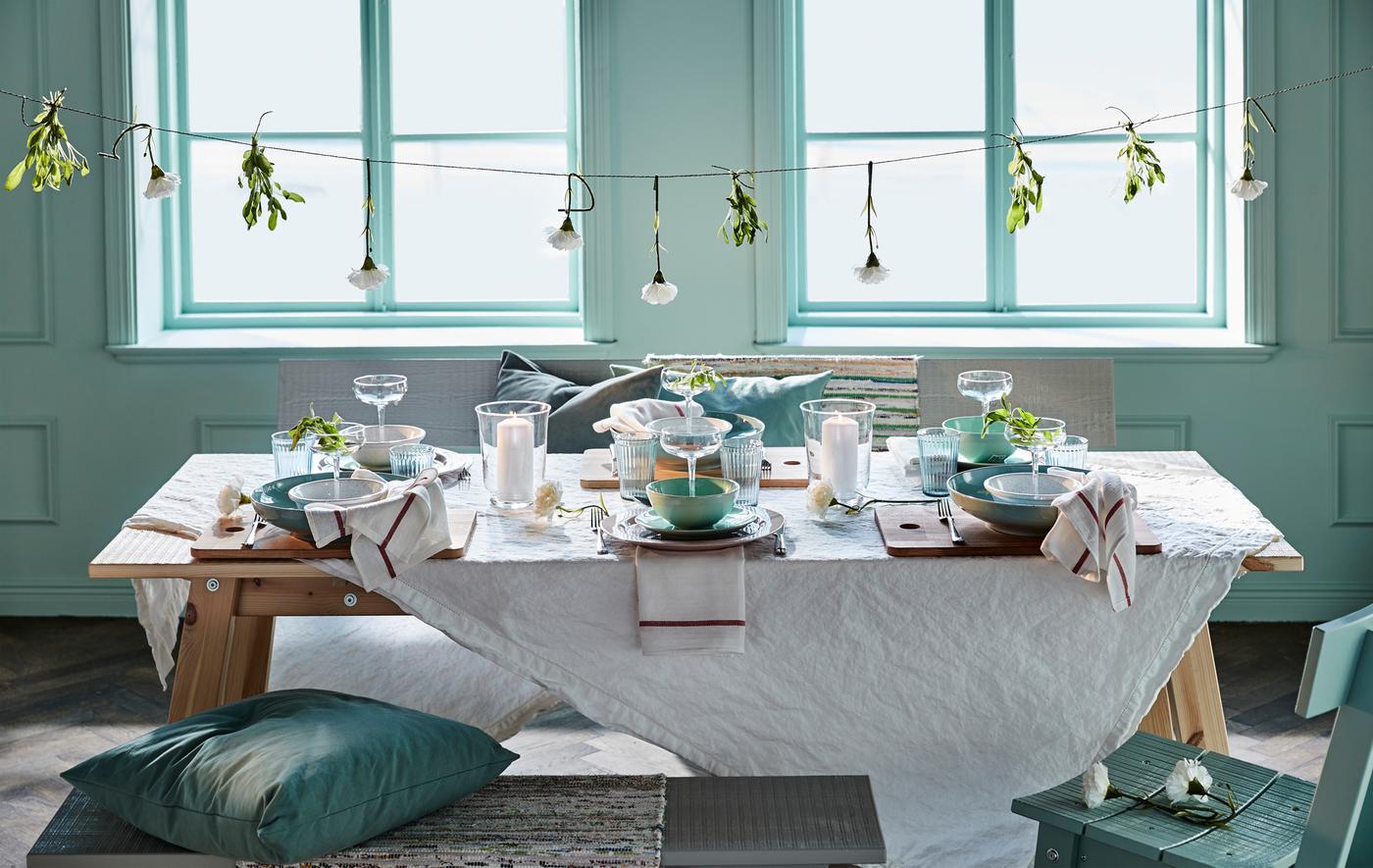 A beautifully set dining table with teal dishware, white linens, candles, and greenery, against a soft blue backdrop with large windows.