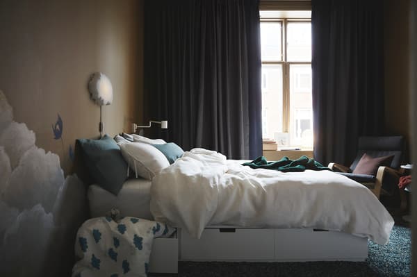 A bed in a dark room with curtains that are slightly open letting a small amount of light in.