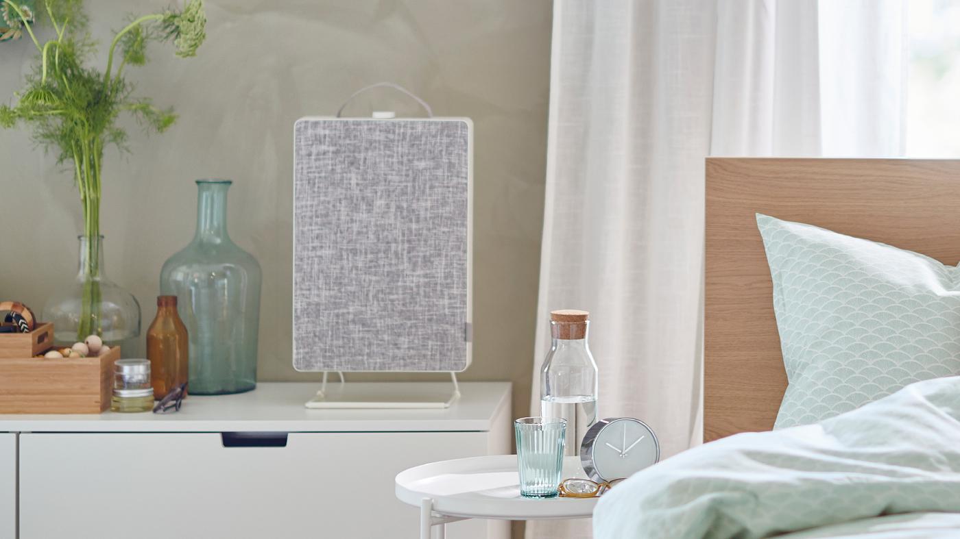 A bed in front of a window and next to a chest of drawers with a FÖRNUFTIG air purifier on top.