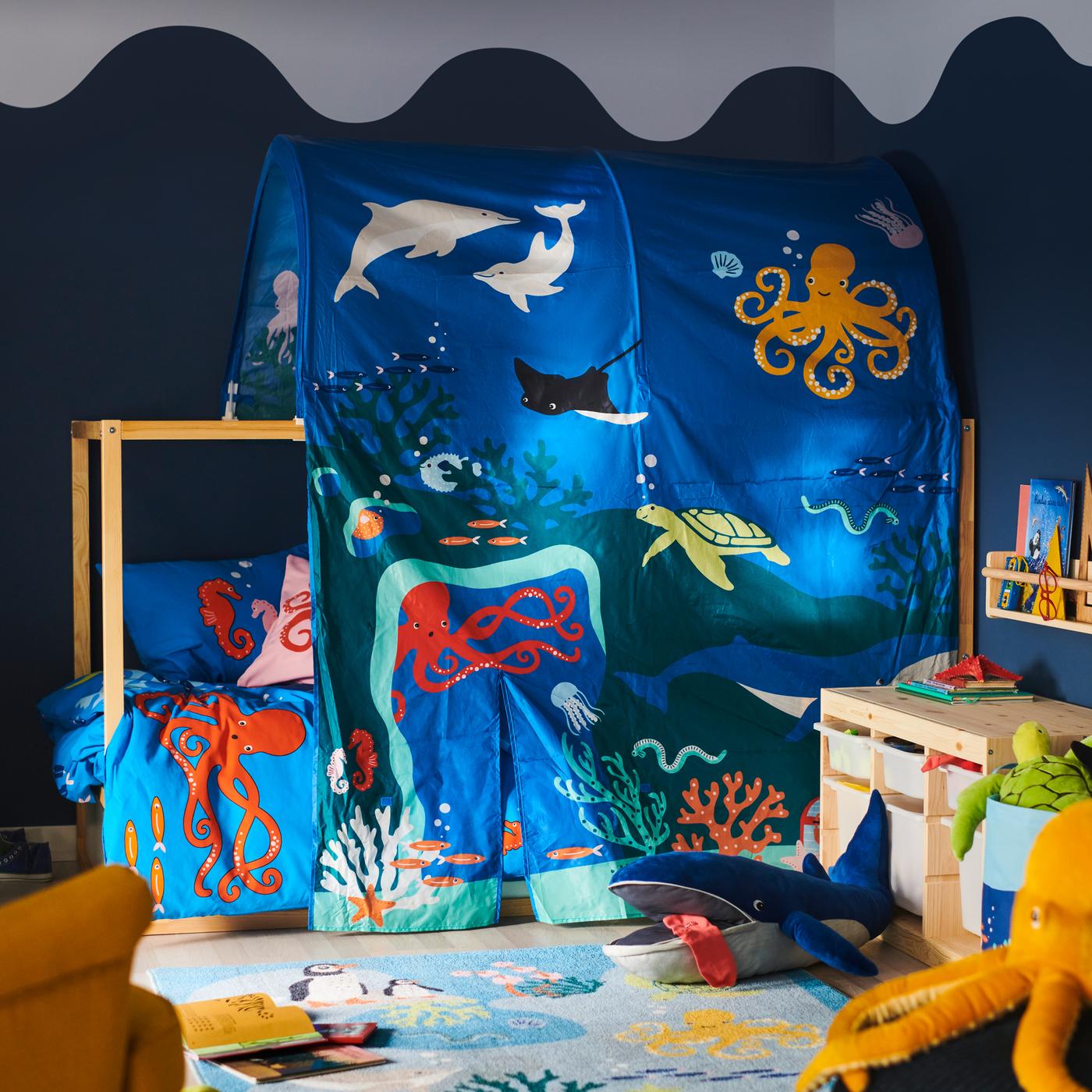 A bed is covered in a KURA ocean animals bed tent; a BLÅVINGAD blue whale soft toy is on a BLÅVINGAD rug in front of it.