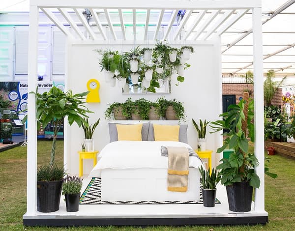 A bed on a platform with white bedding and yellow cushions surrounded bu potted plants.
