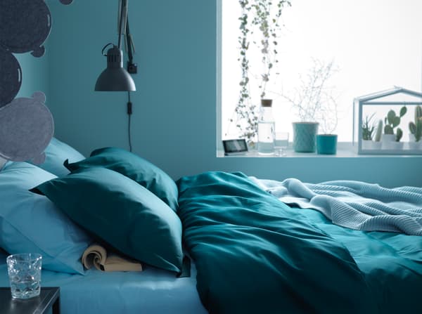 A bed placed by the window and filled with dark green and blue textiles in an overall blue bedroom.