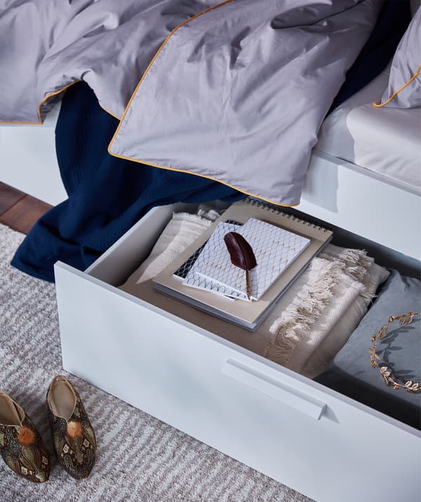 A bed storage box filled with things you might need, under the white BRIMNES bed frame with storage and headboard.