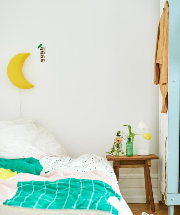 A bed with a green, pink and yellow duvet, a yellow, half-moon shaped light on the wall and wood table with vases beside it.