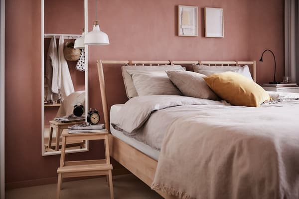 A bed with a wooden frame with footstool side table and full length mirror on a wall.