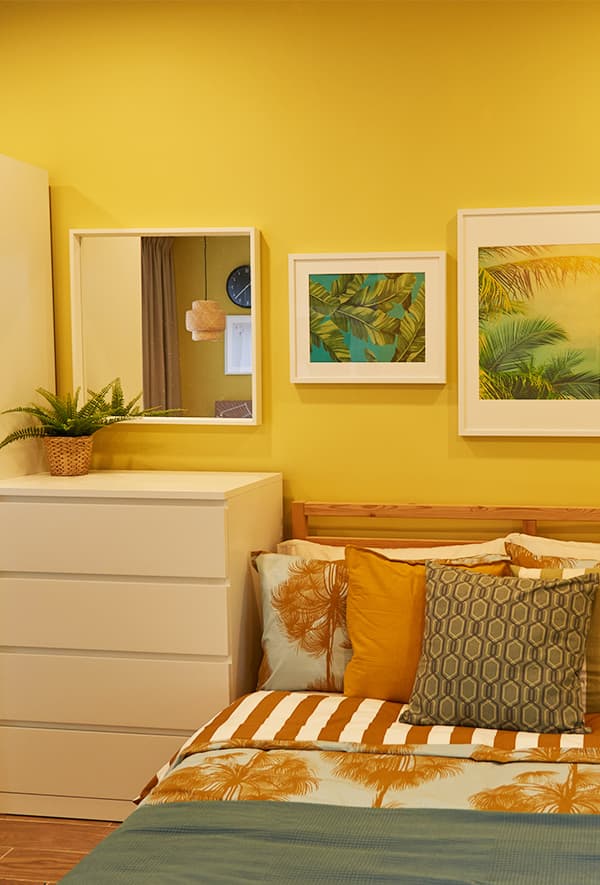 A bed with cushions beside a light birch chest of drawer in a yellow room.