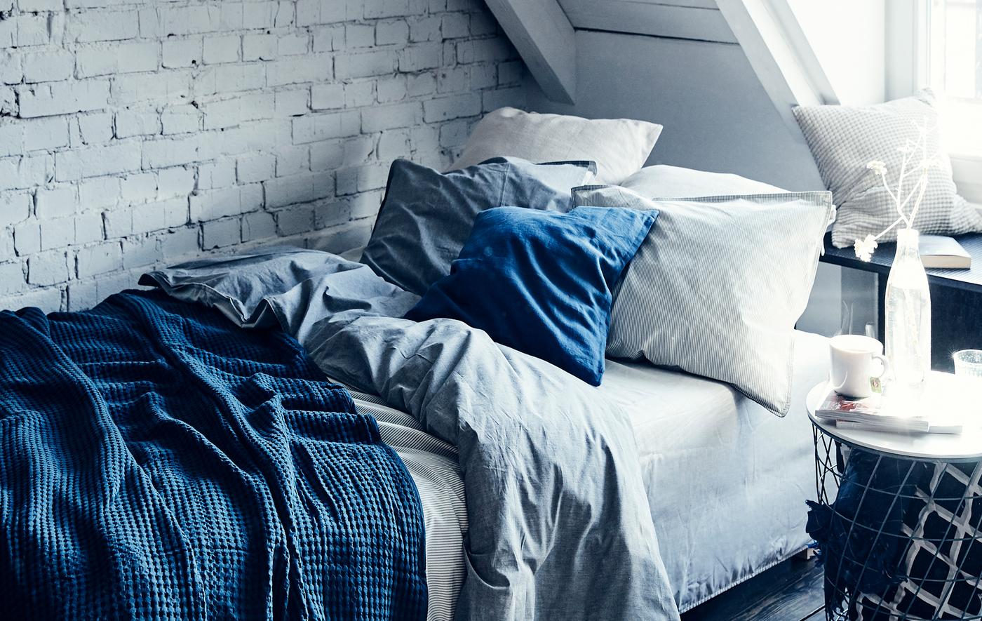 A bed with layered blue and white bedding and cushions against a white painted brick wall.