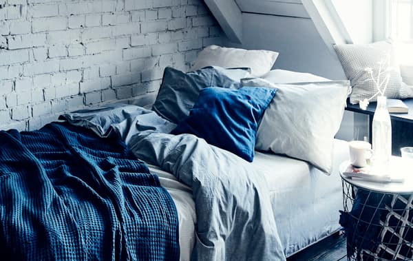 A bed with layered blue and white bedding and cushions against a white painted brick wall