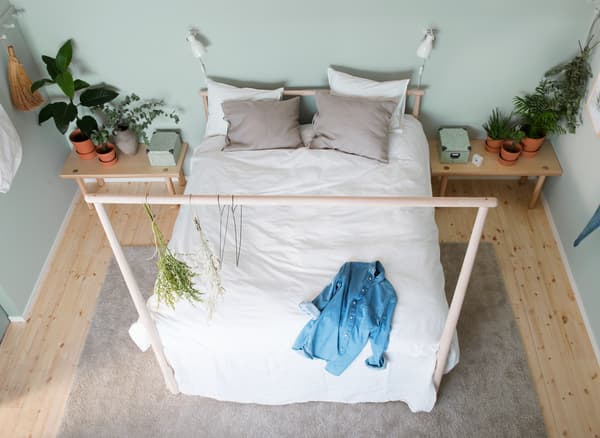 A bed with lightly colored, natural textiles against a light green wall in a symmetrical bedroom with low bedside tables and touches of nature
