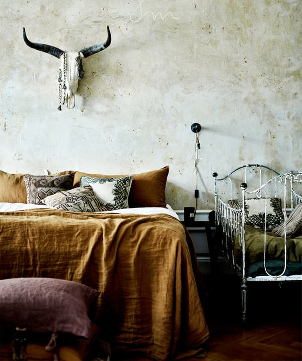 A bed with rust-coloured sheets next to a white metal crib, with an animal skull on the wall above.