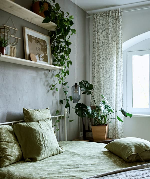 A bedroom decorated with green textiles, plants and two shelves mounted on a grey wall displaying a mix of art and trinkets.
