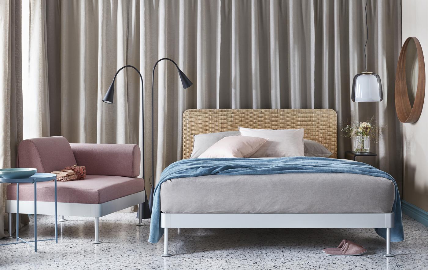 A bedroom in soft neutral grey, beige and pinks featuring a double bed, floor lamp, armchair and side table.