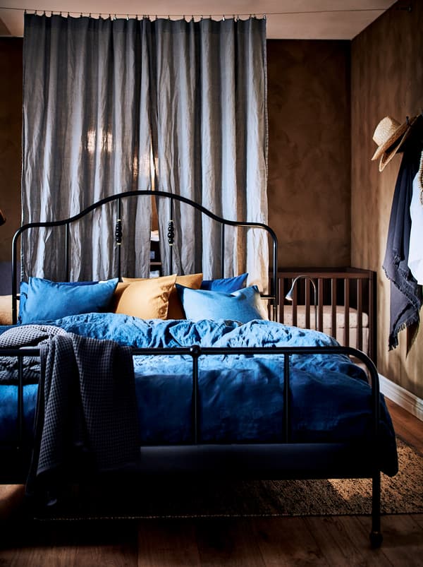 A bedroom slightly divided by two curtains with a black SAGSTUA metal bed frame in the middle of the room, and a baby cot.