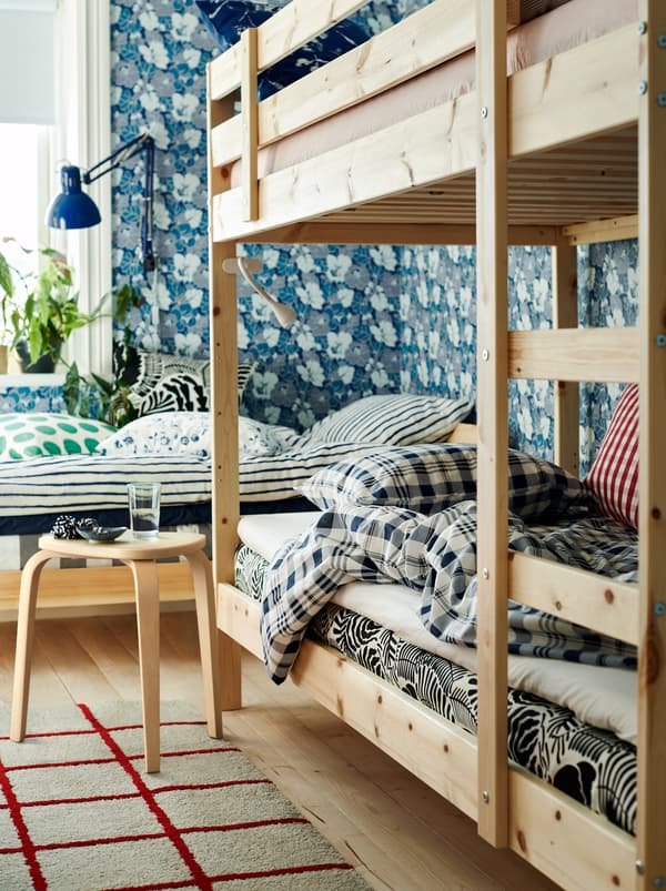 A bedroom with a bed and a bunk bed in neutral colours, a floral wallpaper and textiles in different colours and patterns.