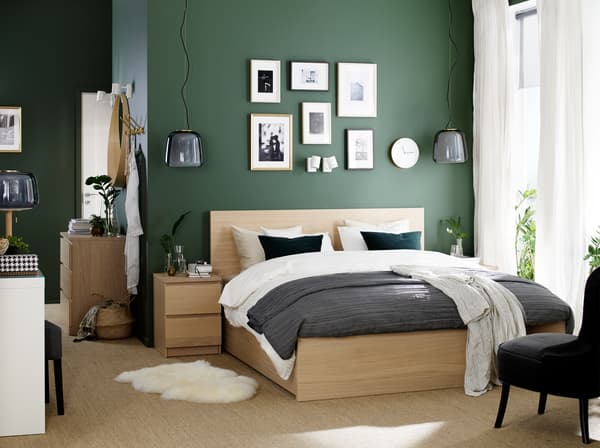A bedroom with a bed frame and bedside table in white stained oak veneer, a white dressing table and a grey armchair.