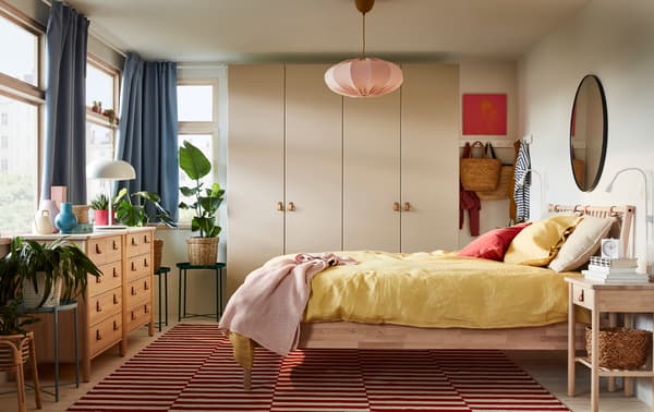 A bedroom with a bed frame and two chests of drawers in birch, a grey-beige wardrobe, blue curtains and yellow bed textiles.