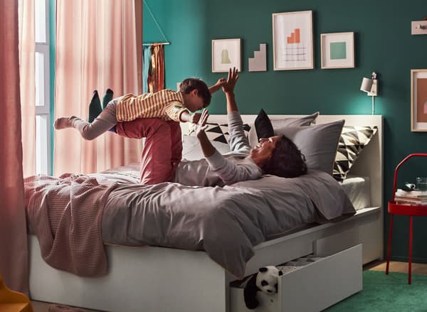 A bedroom with a dad lying on his back with hands and feet up in the air on the bed, playing airplane with his son.