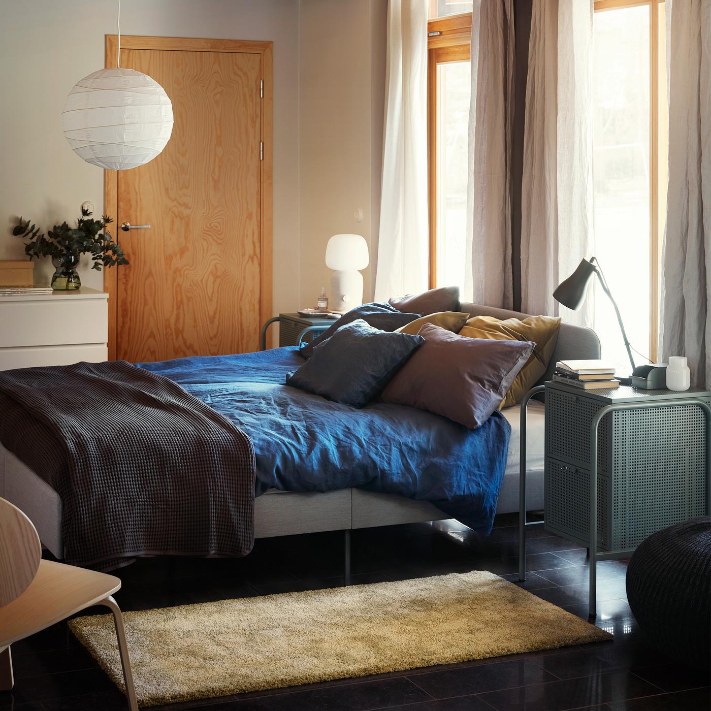 A bedroom with a light SLATTUM upholstered bed frame, blue bed linen, a throw, several pillows and a golden yellow rug.