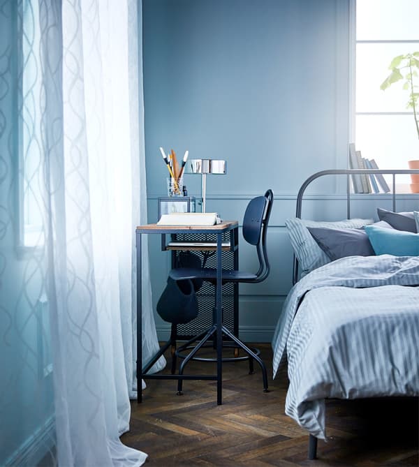 A bedroom with a metal and wood laptop table on castors and a desk chair in the small space between the bed and wall.