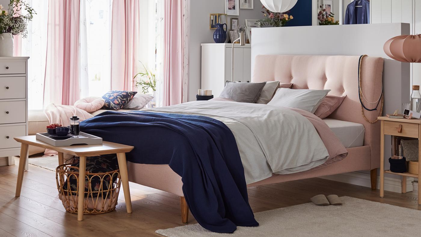 A bedroom with a pink bed with padded headboard, soft coloured textiles, sheer pink curtains and wooden side tables.