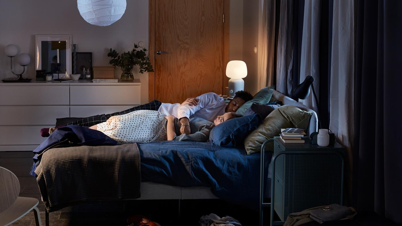A bedroom with a SLATTUM upholstered bed frame, two lamps and dark blue quilt covers, and a couple sleeping on the bed.