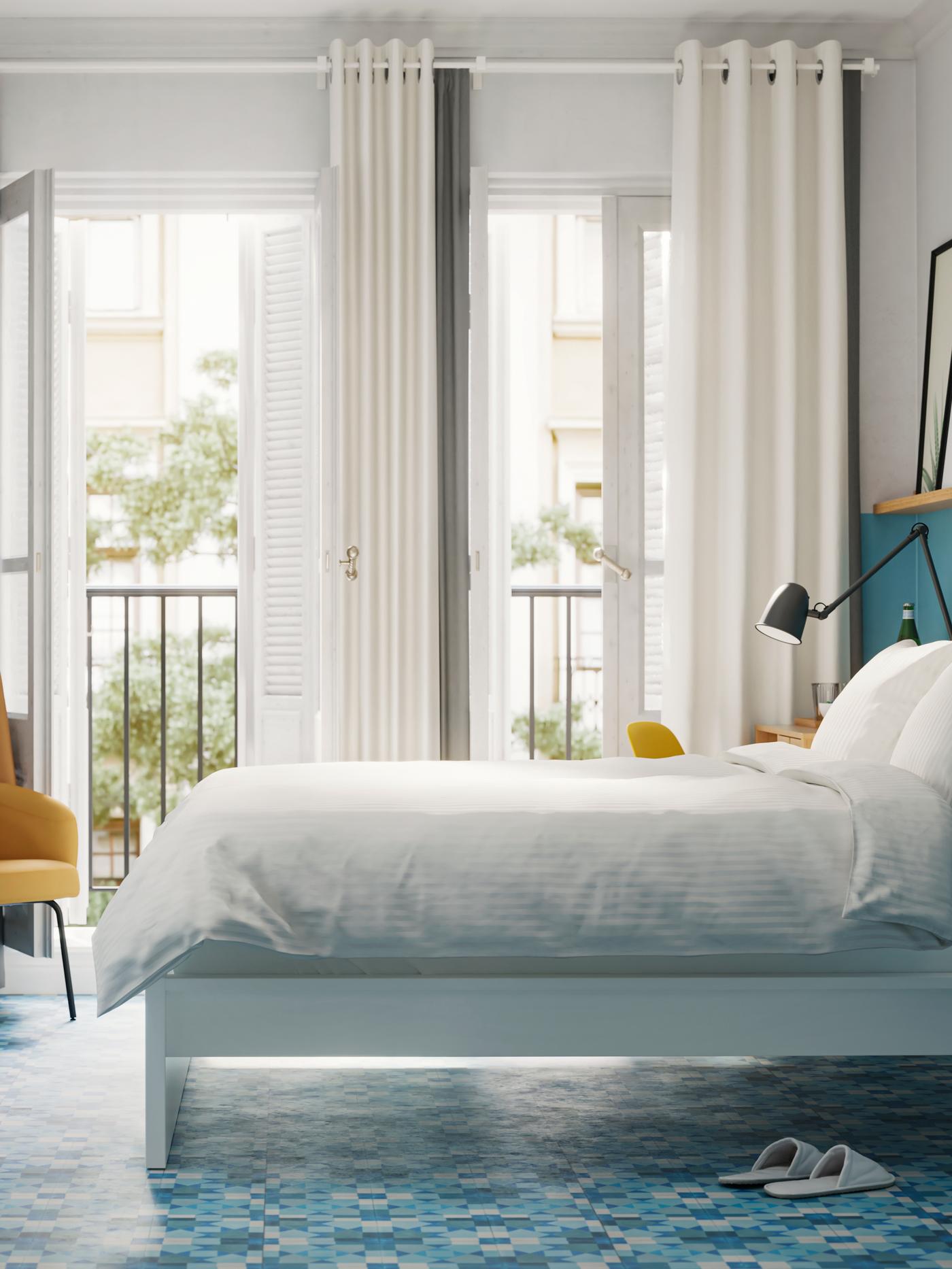 A bedroom with a white bed with white bedding, blue walls and floors and patio doors leading to a balcony.