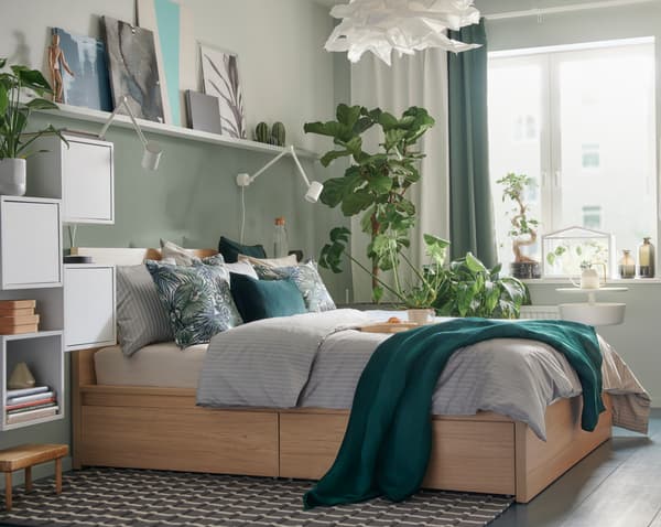 A bedroom with a wood bed, grey and green bedding, picture frames and plants decorating the walls.