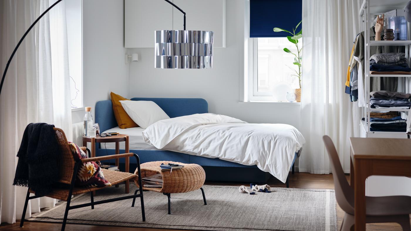 A bedroom with an upholstered bed frame with corner headboard, white quilt cover and pillowcase, a potted plant.