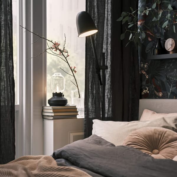 A bedroom with dark curtains hanging and a black lamp shining down on the bed.