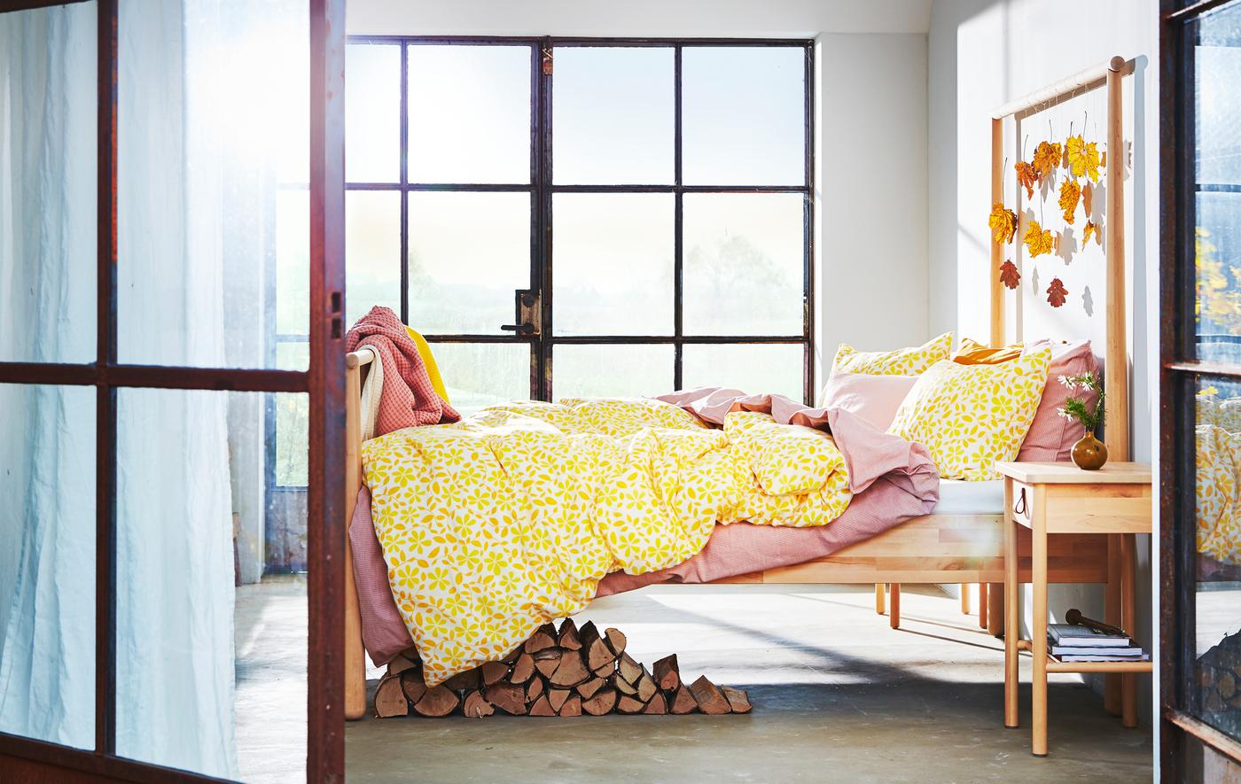 A bedroom with large windows, autumn coloured leaves hanging from a wooden bedframe and yellow and pink layered bedding.
