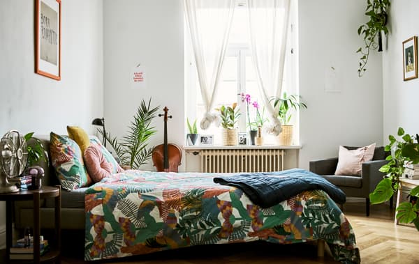 A bedroom with lots of plants, sheer curtains at the window, parquet flooring and a bed covered with a tropical-print duvet.