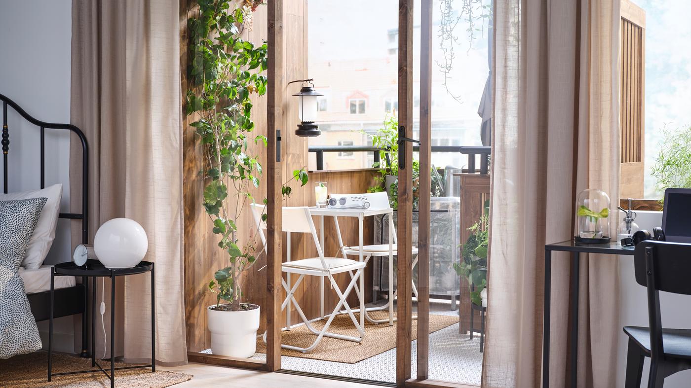 A bedroom with open doors to the balcony that has a white FEJAN table and two chairs on it and lots of green plants.