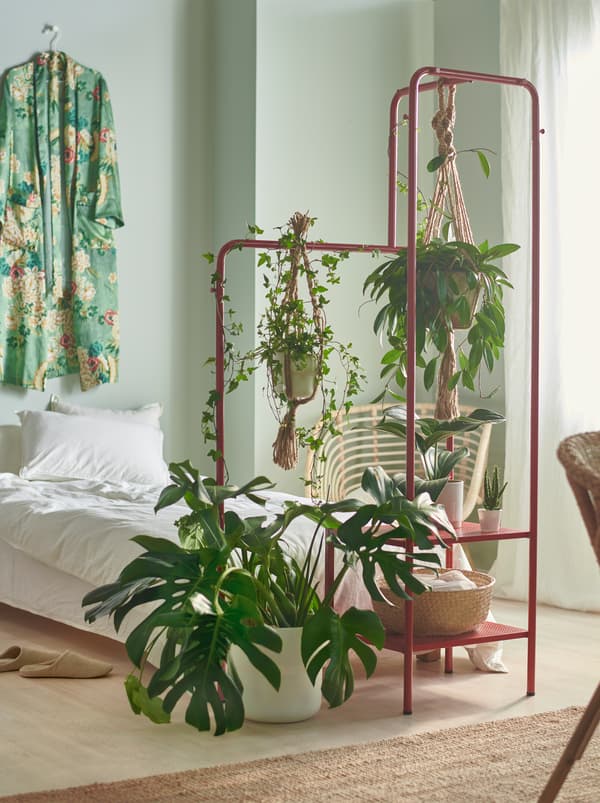 A bedroom with small and large plants at the end of the bed, hanging and standing on a red NIKKEBY clothes rack.