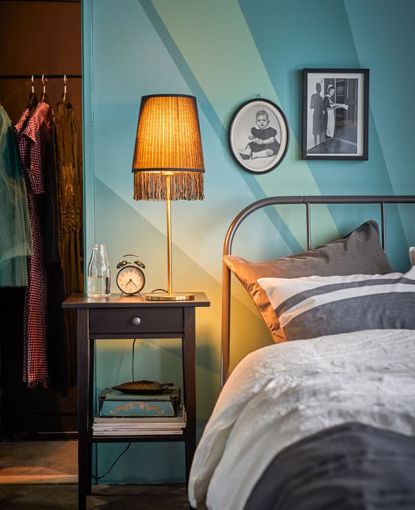 A bedside table with a gold, fring lamp sits next to a bed.