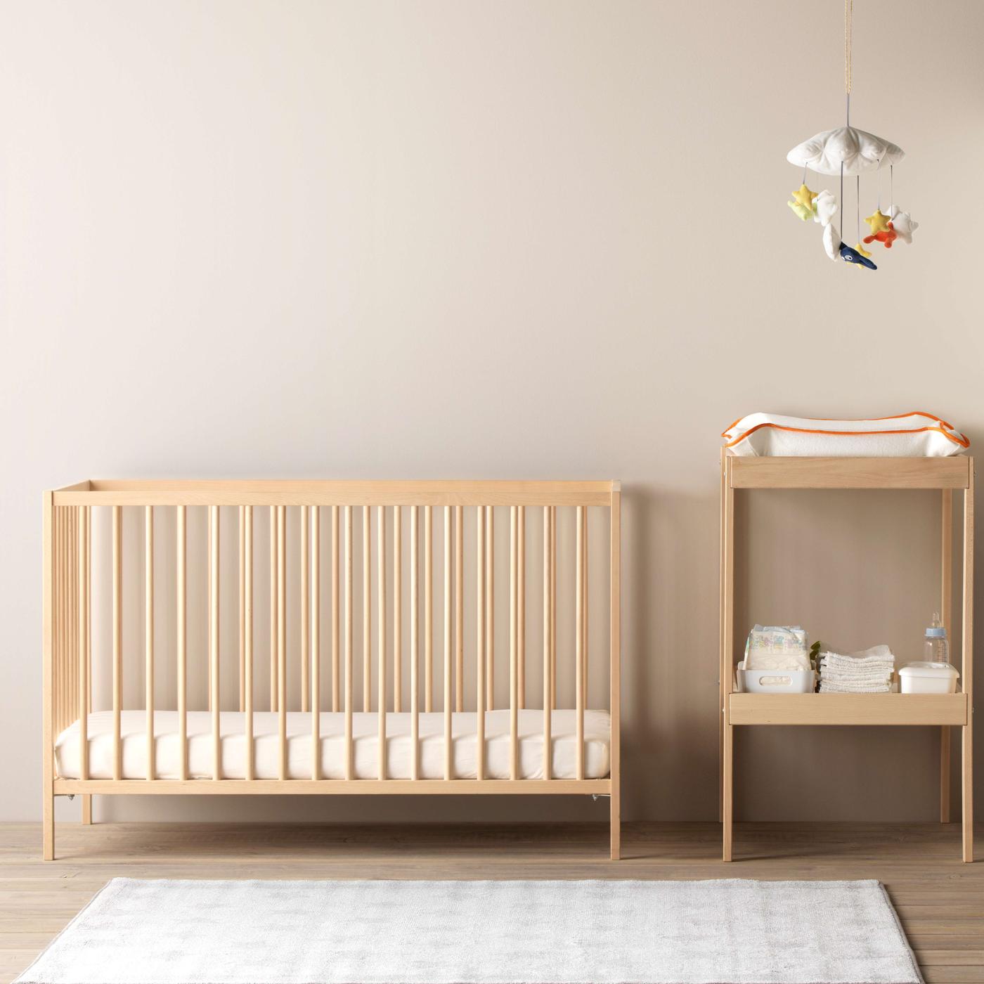 A beech SNIGLAR crib with only a mattress and fitted sheet in it, while a changing table is next to it with a  GULLIGAST mobile that hangs high above it.