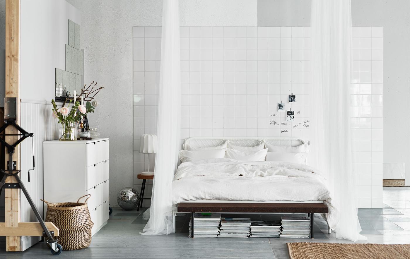 A big white bed low to the floor with crisp white sheets and pillows and sheer white curtains that drape to the floor.
