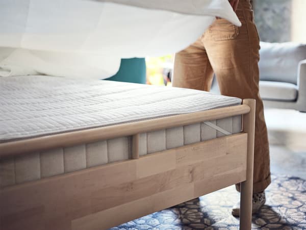 A birch bedframe with a thick beige mattress and white mattress protector. A man is making the bed with white bedding.