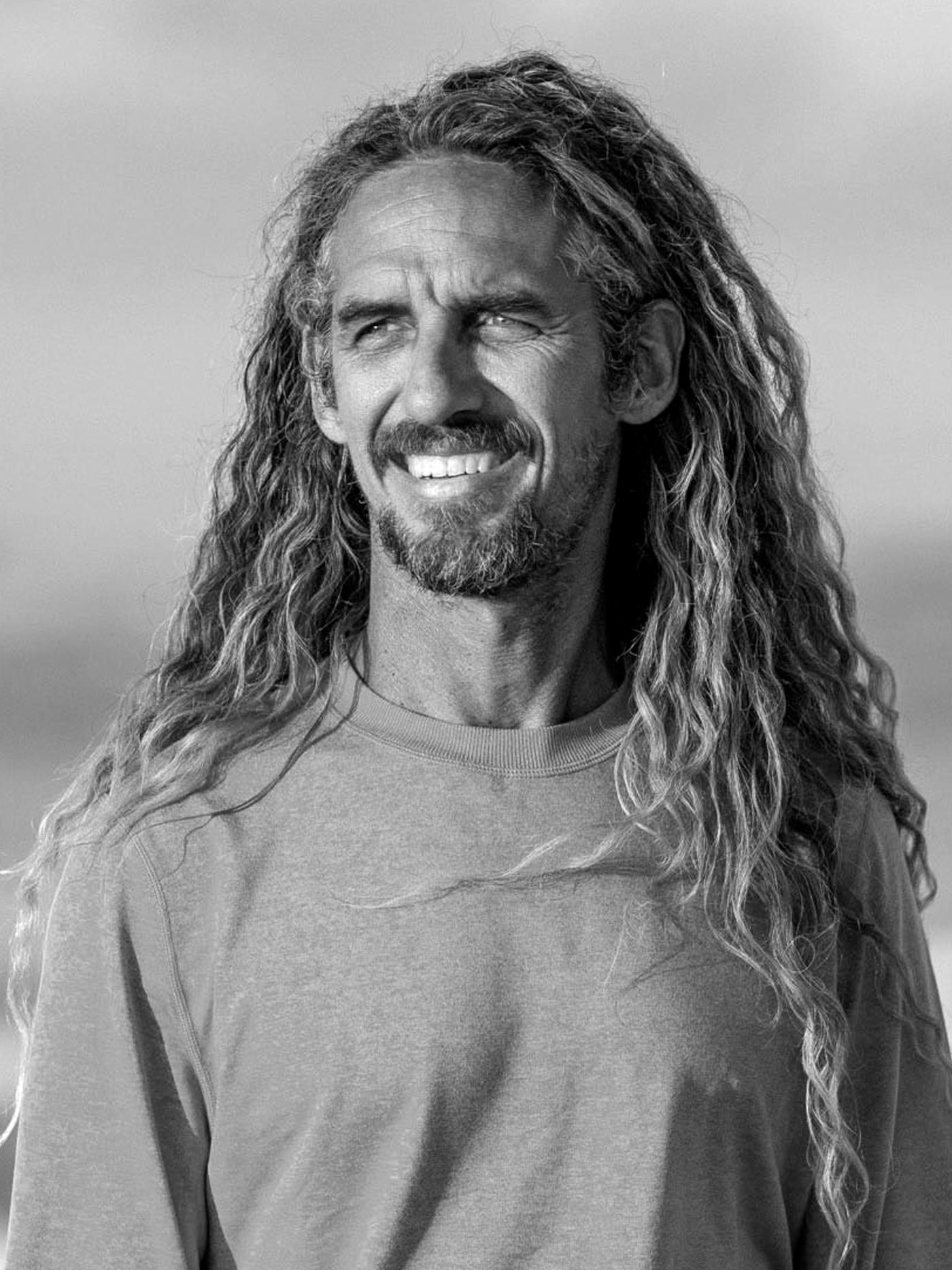 A black and white photograph featuring the former pro-surfer Rob Machado wearing a t-shirt, smiling by the sea.