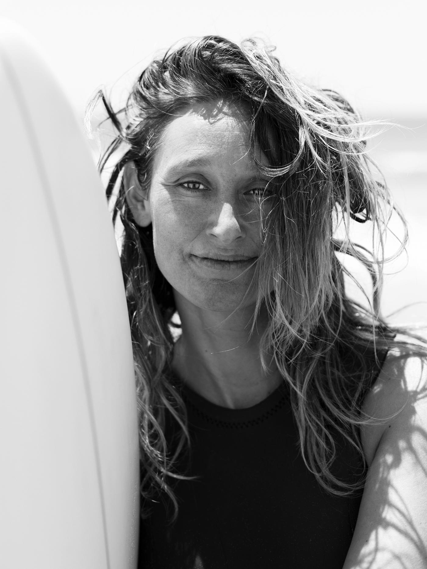 A black and white photograph featuring the former pro-surfer Kassia Meador standing by a white surfboard by the sea.
