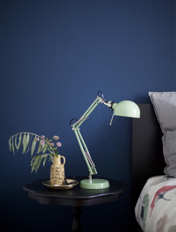 A black bedside table and mint green lamp against a dark blue wall.