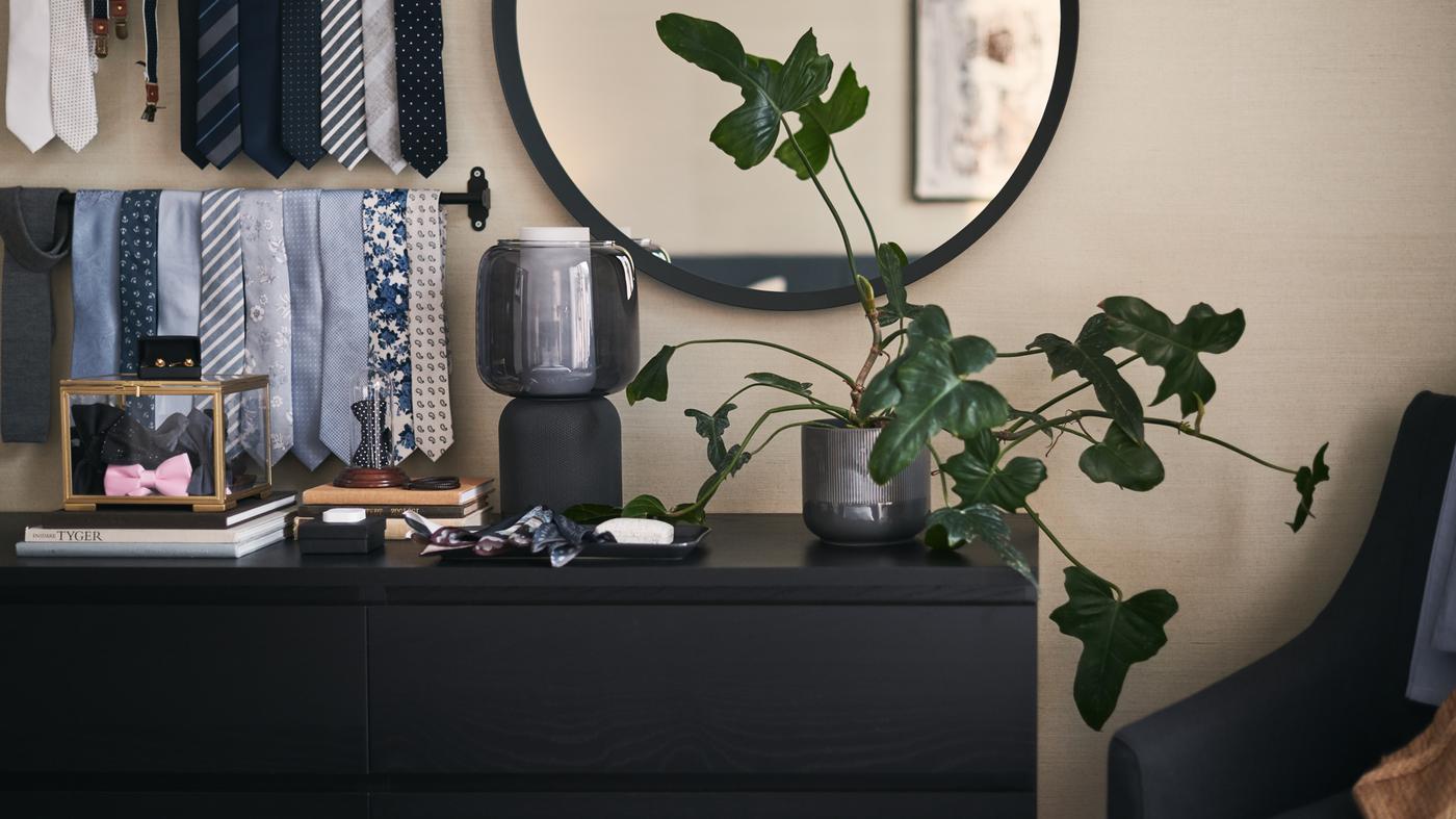 A black-brown MALM chest of drawers in front of a large round mirror with a leafy plant and SYMFONISK WiFi speaker on top.